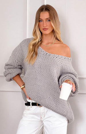 blonde model wearing a grey oversized chunky knit sweater with an asymmetric neckline paired with white jeans