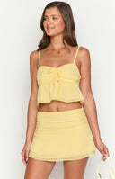 Brunette girl wearing a yellow two piece set, flowy crop top and mini skirt with a gold flower necklace and holding a drink.